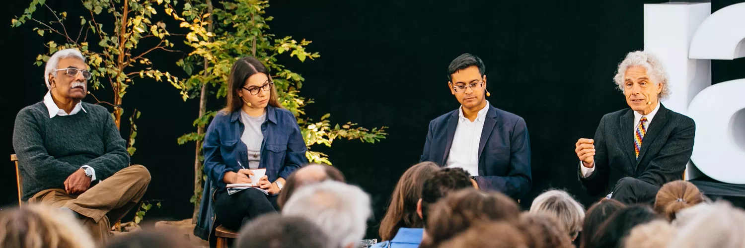 A panel discussion featuring four individuals, with two men and one woman engaged in conversation, surrounded by greenery and an attentive audience.