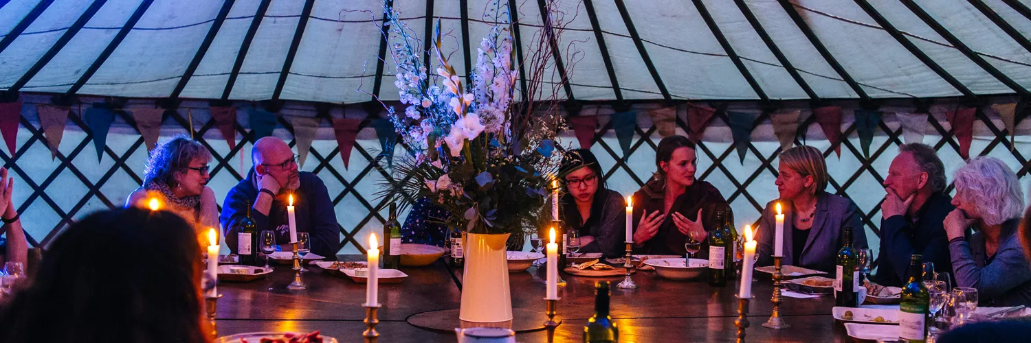 A group of elegantly dressed people engaged in conversation at a candlelit dinner inside a yurt, surrounded by floral arrangements and festive decorations.