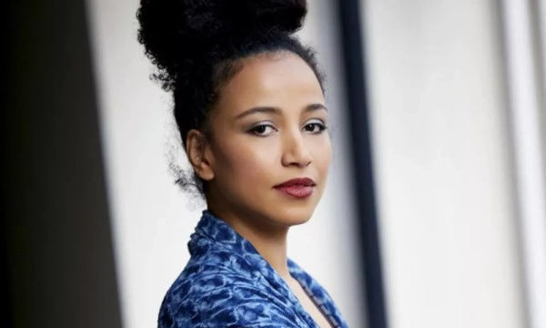 A woman with curly hair styled in a bun, wearing a blue patterned blouse, gazes thoughtfully towards the camera with a natural makeup look.