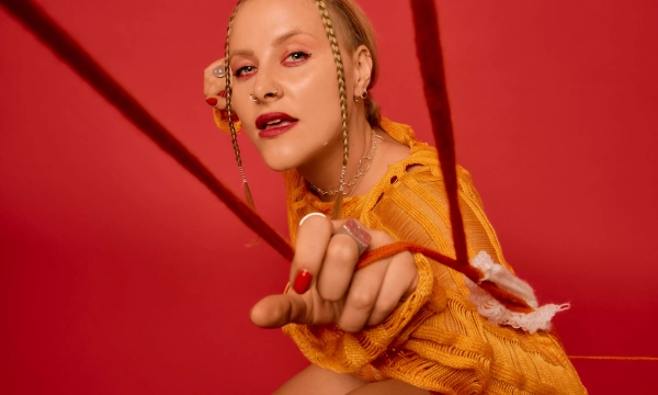 A person with braided hair is striking a pose against a vivid red background, wearing an orange knit sweater and high-heeled boots, creating a bold fashion statement.
