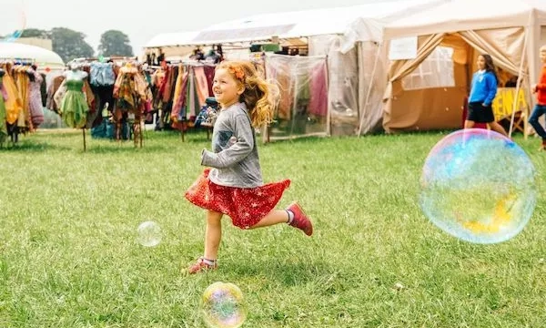 A young girl with red hair joyfully runs through a grassy area at a festive outdoor market, surrounded by colorful clothing tents and large bubbles.