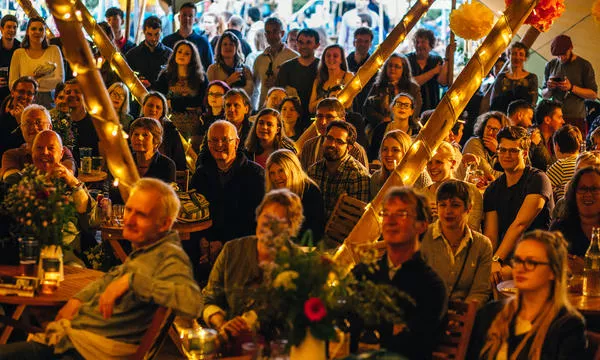 A lively crowd gathered in a festive tent, filled with people enjoying an event, decorated with lights and flowers, and displaying a warm, cheerful atmosphere.