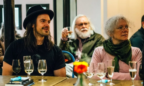 A group of three people sitting at a table with glasses of drinks, including a man with long hair and a hat, a woman with curly gray hair and a scarf, and a man with a white beard in the background.