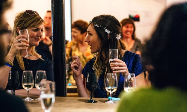 Two women engaged in conversation at a social gathering, holding glasses of wine and champagne, with others blurred in the background.