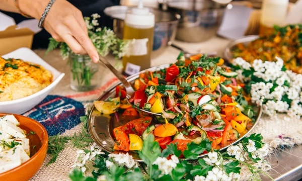 A colorful, fresh salad is being served from a large platter, surrounded by herbs and garnishes, with bowls of other food items displayed in the background.