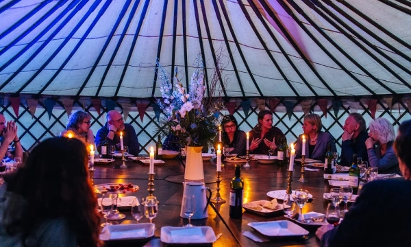 A group of people dining around a large circular table inside a yurt, illuminated by candles and colorful lights, with flowers as a centerpiece.