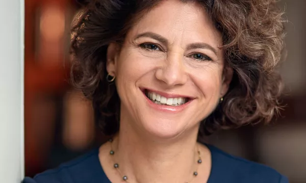 Smiling woman with curly hair, wearing a dark blue top and a necklace, stands against a softly lit background.