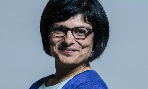 A woman with short black hair and glasses, wearing a blue cardigan, smiles softly against a gray background.