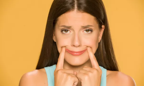 A woman with long brown hair wears a light blue top, pulling a forced smile with her fingers against a yellow background.