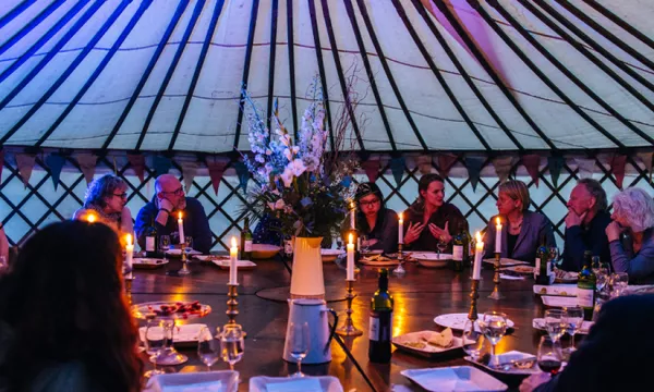 A group of people seated around a large circular table inside a yurt, adorned with candles and flowers, engaged in conversation during a dinner event.