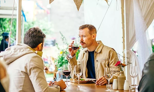 Two men dining at an outdoor table, one enjoying a glass of red wine while the other focuses on his meal. Decor includes flowers and festive decorations in the background.