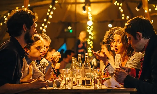 A lively dinner scene featuring a diverse group of people engaged in animated conversation at a long table, illuminated by warm string lights in a cozy setting.