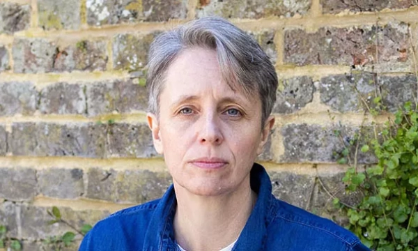 A person with short gray hair, wearing a denim jacket, stands in front of a stone wall with greenery.