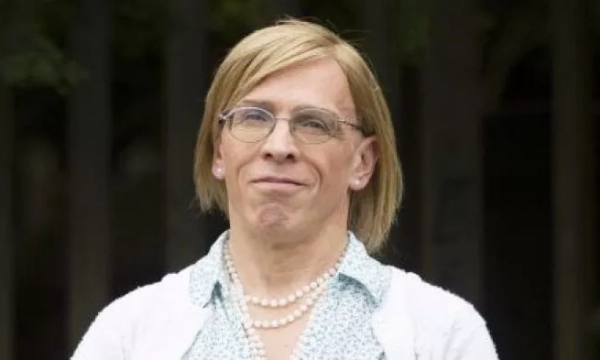 A person with shoulder-length blonde hair, wearing glasses, a white blouse, and a pearl necklace, stands outdoors with a neutral expression.