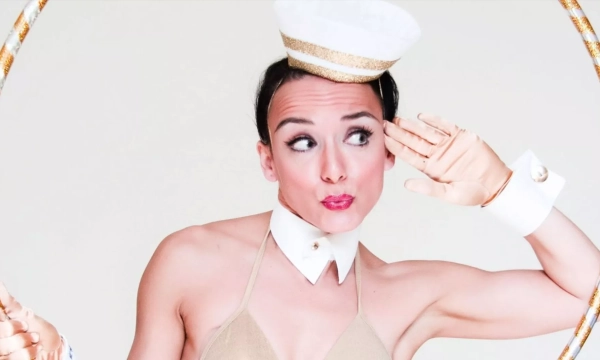 A performer in a gold bikini and sailor hat playfully poses with a hula hoop, giving a cheeky salute with an exaggerated expression.