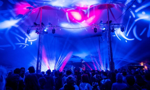 A vibrant concert scene inside a tent, with colorful blue and pink lighting creating dynamic patterns above a crowd of people enjoying the performance.