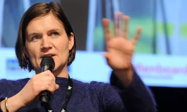 A woman with short hair speaks into a microphone while gesturing with her hand. She appears to be participating in a discussion or panel event.