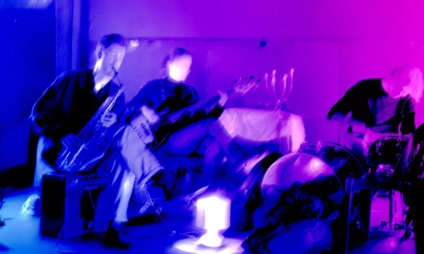 Two musicians play instruments in a dimly lit room, with vibrant purple lighting. One plays a saxophone, while another is on bass, and a drummer is in the background.