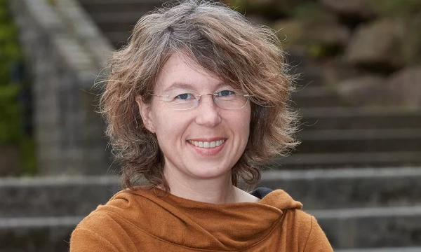 A woman with curly hair wearing glasses and an orange sweater smiles in front of a stone staircase and greenery.