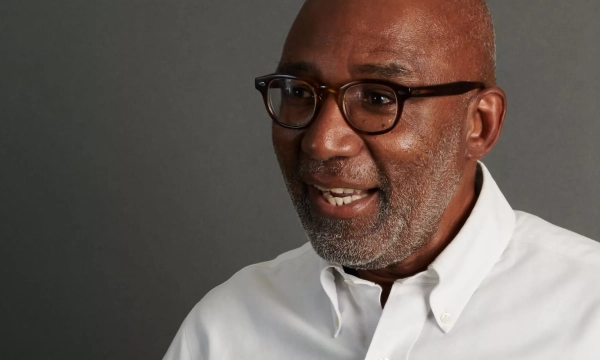A smiling man with glasses and a short beard, wearing a white shirt, engaged in conversation against a gray background.