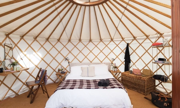 A cozy yurt interior featuring a bed with blankets, wooden furniture, and decorative items, all under a circular fabric ceiling.