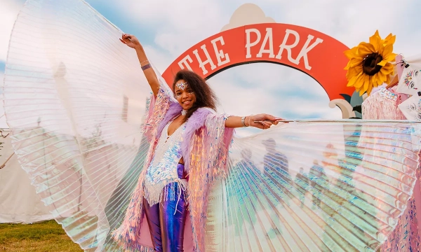 A person in colorful attire with shimmering wings stands joyfully in front of a whimsical sign reading 