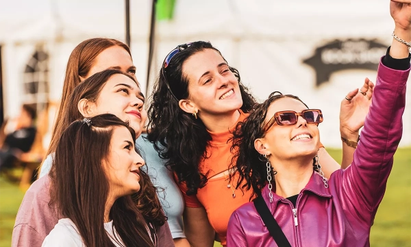 A group of five women smiling and posing for a selfie outdoors, wearing stylish clothing and sunglasses, with a tent in the background.