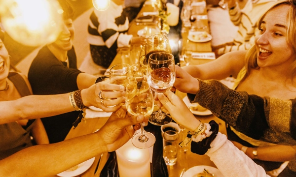 A joyful gathering with diverse people toasting with drinks around a dinner table, celebrating a special occasion with laughter and warm lighting.