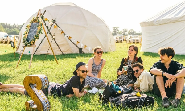 A group of five friends relaxes on the grass in a festival setting, with white tents in the background and decorative items nearby.