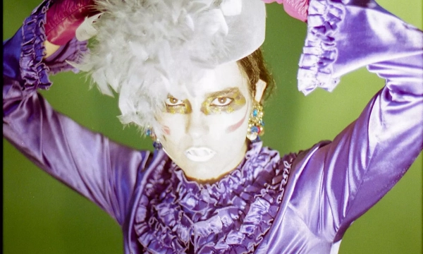 A person with dramatic makeup and a stylish outfit poses against a bright green background, showcasing bold fashion and theatrical flair.
