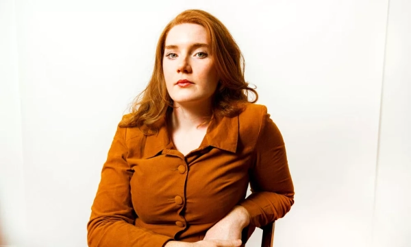 A woman with long, wavy red hair sits against a plain white background, wearing a brown button-up blazer and a serious expression.