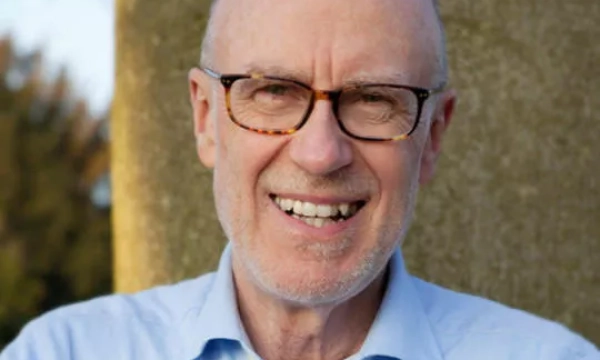 A smiling older man with glasses, wearing a light blue shirt, stands outside in front of a stone background, surrounded by greenery.