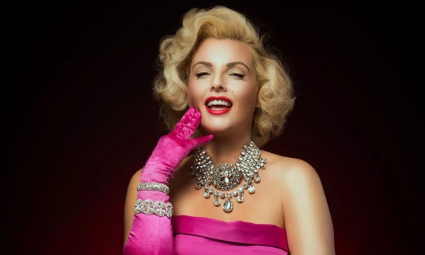 A glamorous woman with blonde curls, wearing a pink off-the-shoulder gown and vibrant gloves, adorned with a sparkling necklace, smiles confidently against a dark background.
