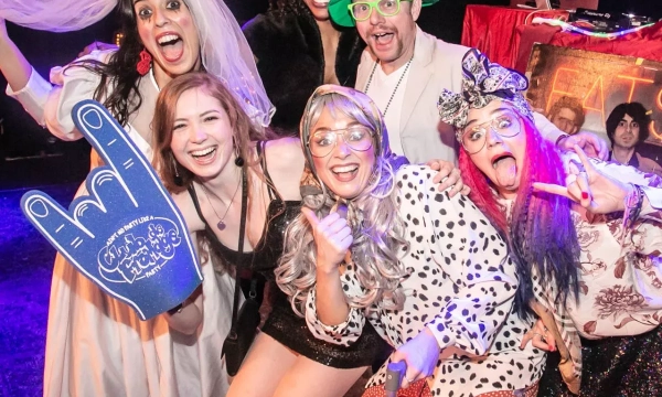 A lively group of six people in colorful costumes and accessories, celebrating with playful poses and expressions at a music event.