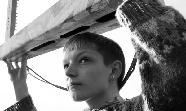 A young person in a patterned sweater holding a wooden instrument above their head, set against a bright sky.