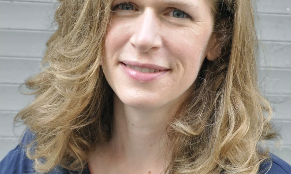 A woman with wavy, shoulder-length hair poses for the camera, wearing a navy blue top, smiling softly against a gray background.