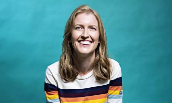 Smiling woman with long blonde hair wearing a striped sweater, against a bright turquoise background.