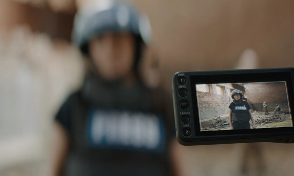 A journalist wearing a bulletproof vest and helmet stands in a war-torn area, while being filmed by a camera displaying their image on the screen.