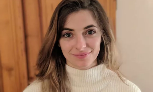 A young woman with long brown hair wearing a white turtleneck sweater, standing against a wooden background.