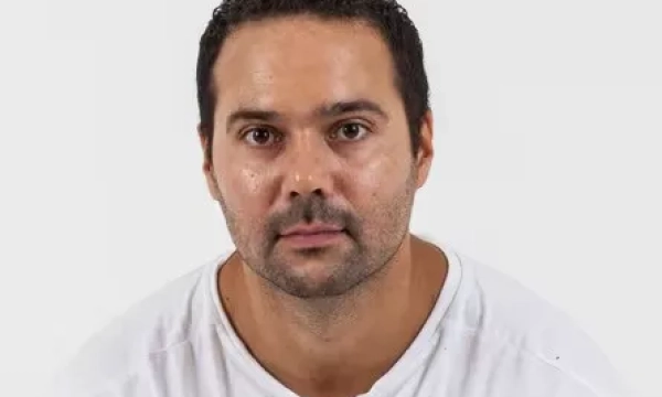 A man with short dark hair, wearing a plain white shirt, has a serious expression while facing the camera against a plain white background.