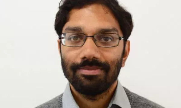 A man with a beard and glasses, wearing a gray sweater over a collared shirt, stands against a plain white background.