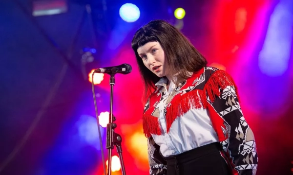 A singer with dark hair and a fringe wearing a fringed red and black patterned shirt performs on stage, illuminated by colorful stage lights.