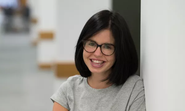 Smiling woman with short black hair and glasses, wearing a gray shirt, leaning against a white wall in a well-lit interior space.