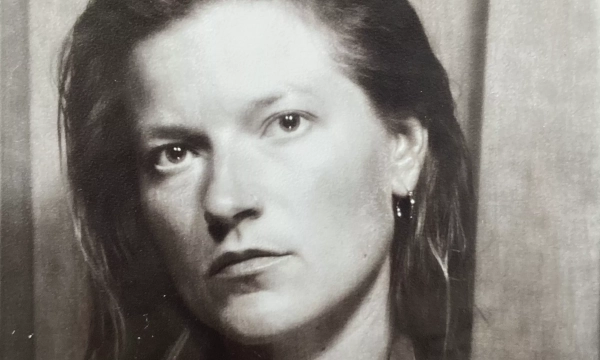 A black-and-white portrait of a woman with long hair, wearing a jacket, looking serious and contemplative against a textured backdrop.