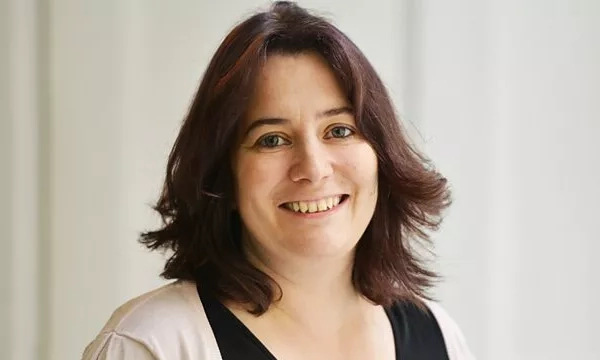 A woman with shoulder-length dark hair smiles warmly at the camera, wearing a light-colored top against a softly lit background.
