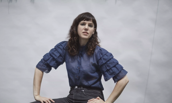 A woman with dark hair styled in loose waves poses confidently in a blue ruffled blouse and high-waisted black jeans against a textured white backdrop.