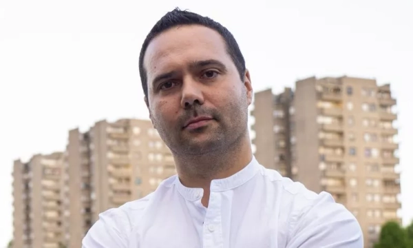 A man with a serious expression stands with arms crossed, wearing a white shirt, against a backdrop of residential buildings.
