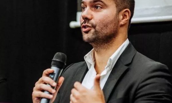 A man in a black suit holds a microphone while speaking at a panel discussion, with drinks in red cups visible in the foreground.