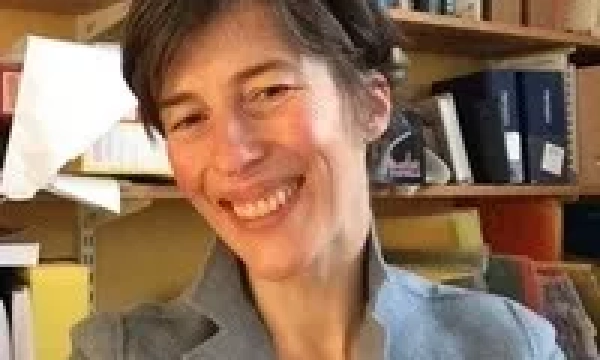 A smiling person with short, tousled hair wearing a gray blazer, seated in front of a bookshelf filled with books and various items.