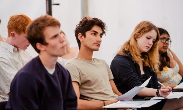 A diverse group of students listen attentively during a lecture or discussion, seated in a tent with notes and papers in hand.